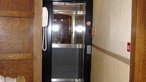 A hotel elevator with a red carpet leading to it, a phone on the wall, and a wooden panel design on the wall. - Hotel Ensor (Bruges)