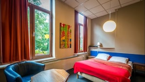 A hotel room with a bed, a chair, a table, a window with curtains, a radiator, a wall-mounted lamp, and a colorful painting. - Hotel Ensor (Bruges)