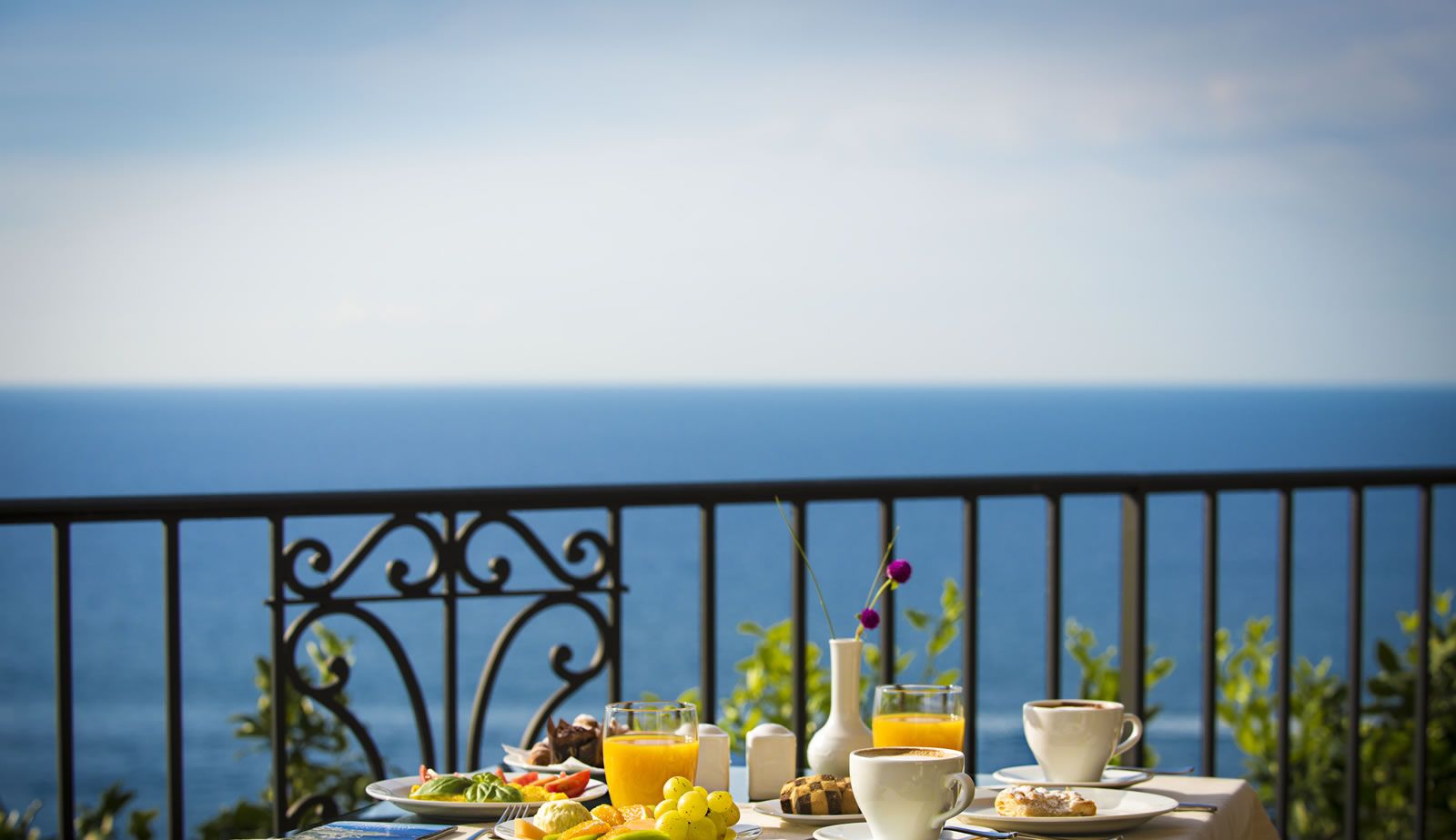 A table set with breakfast items, including fruits, pastries, and beverages, overlooking a scenic ocean view.