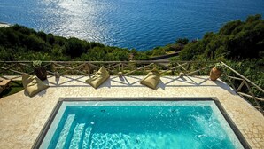 Piscina rectangular al aire libre con agua azul cristalina, rodeada por una terraza de piedra y una pérgola de madera con cojines.