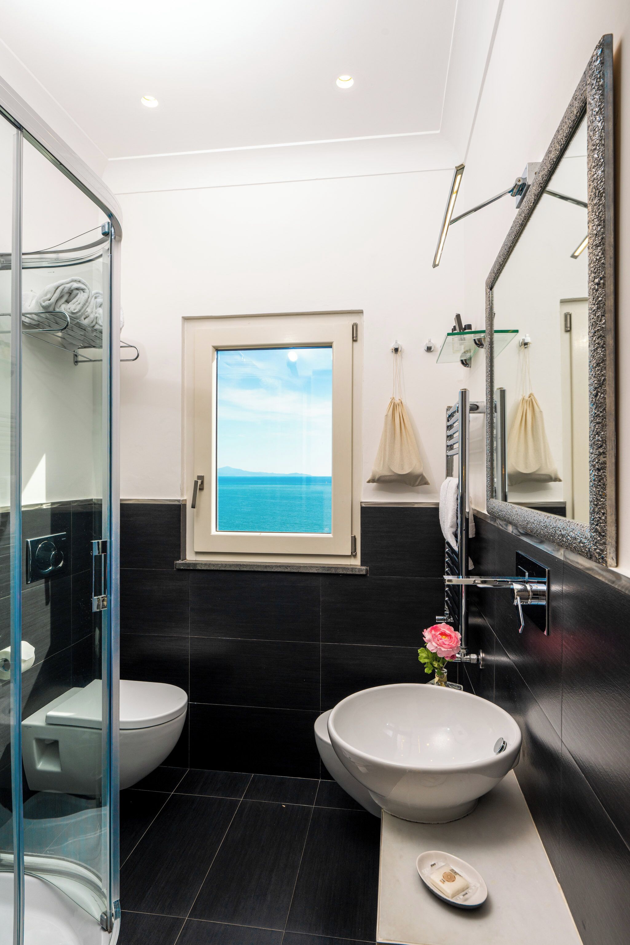 Un baño moderno con ducha de vidrio, un amplio ventanal con vista al mar, un lavabo blanco y un espejo con marco texturizado.