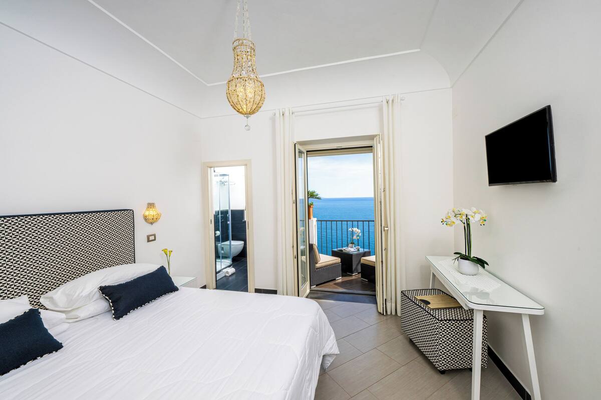Een hotelkamer met een bed, bureau, televisie en balkon met uitzicht op zee.