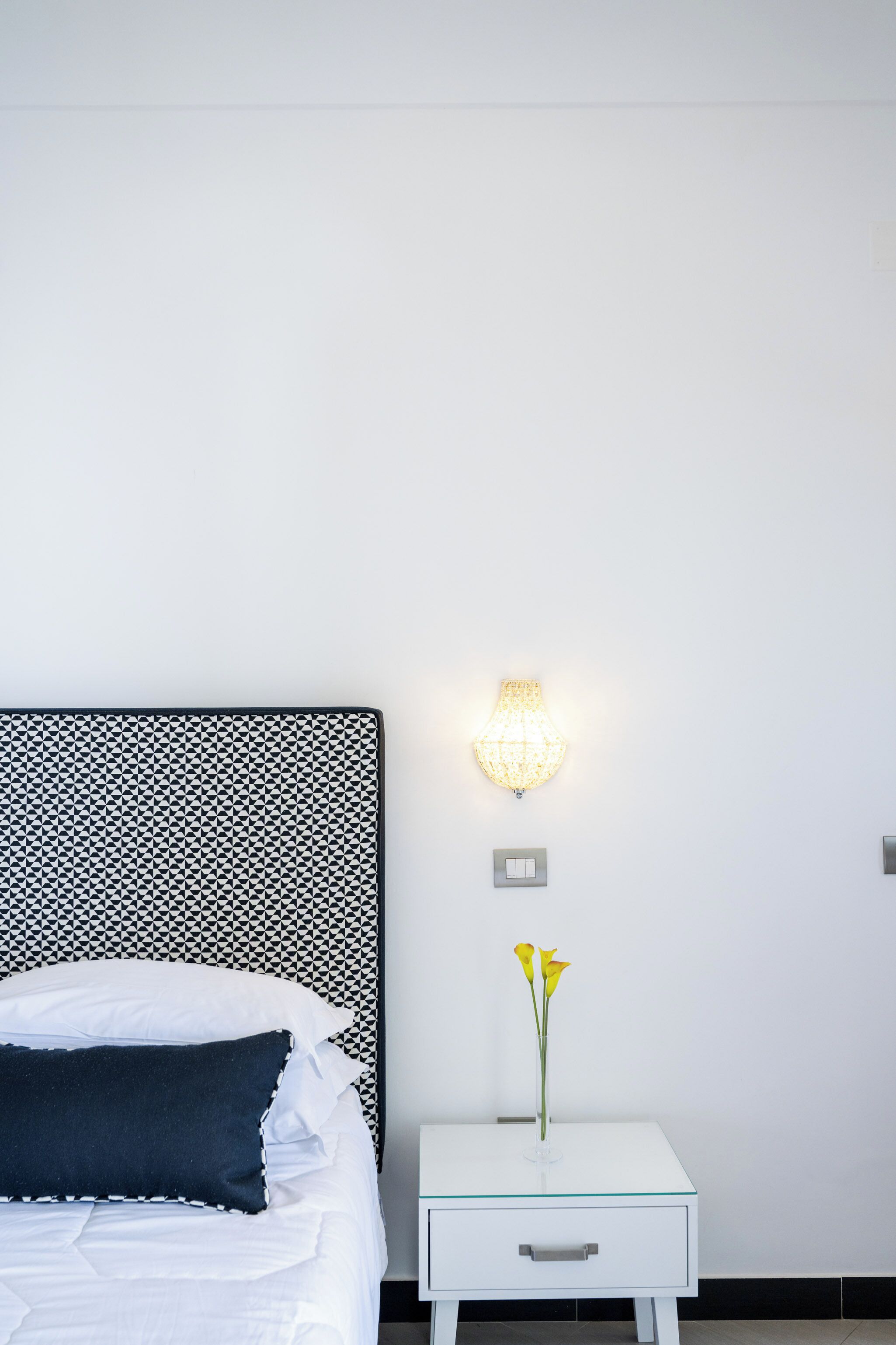 Cama bem arrumada com almofadas brancas e escuras, mesa de cabeceira com tampo de vidro e luminária fixada na parede.