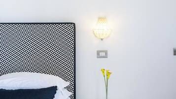 Cama bem arrumada com almofadas brancas e escuras, mesa de cabeceira com tampo de vidro e luminária fixada na parede.