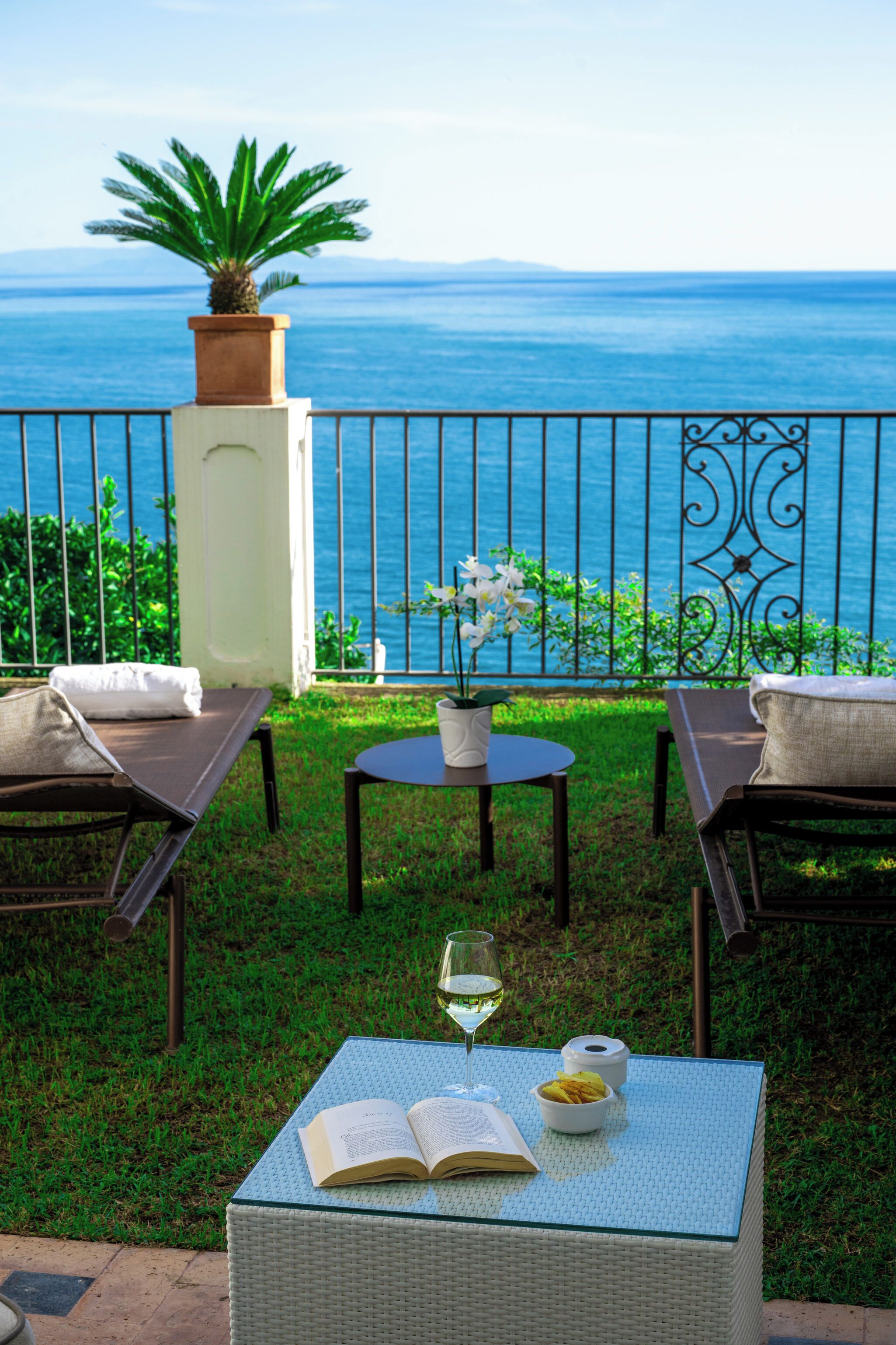 A terrace with a view of the sea, featuring a wicker table with a book, a glass of wine, and a bowl of fruit.