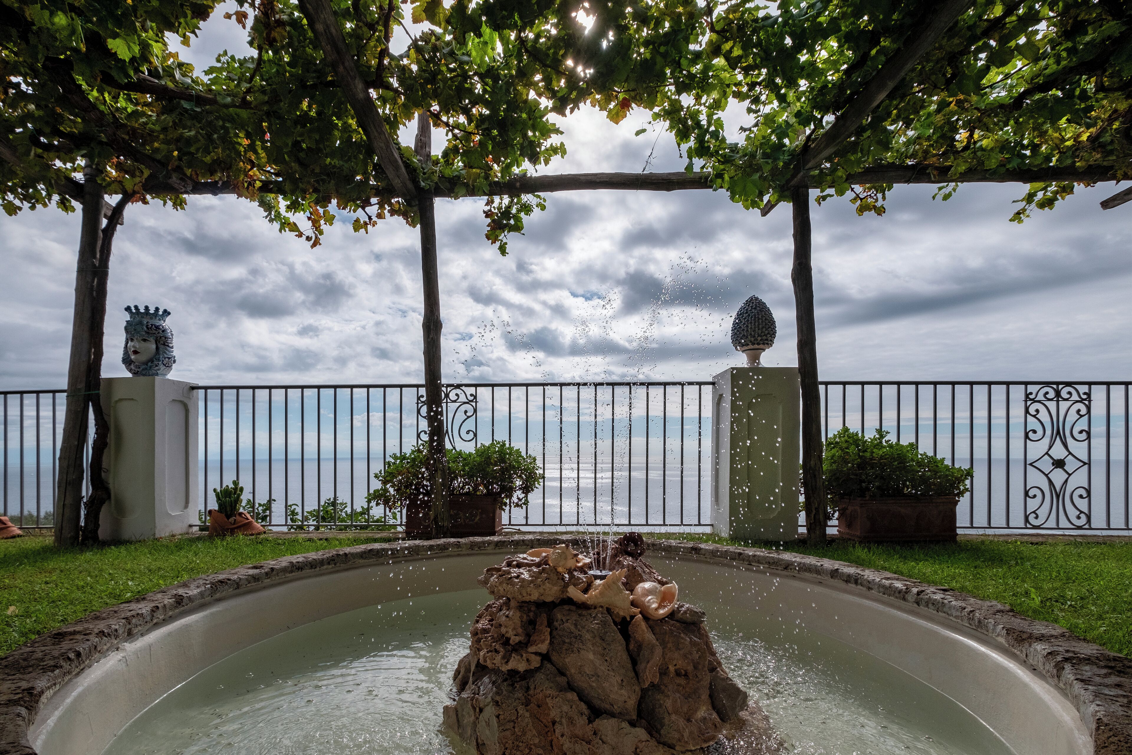 A fountain with water spouting from a rock formation, surrounded by a circular pool and a pergola with a view of the sea.