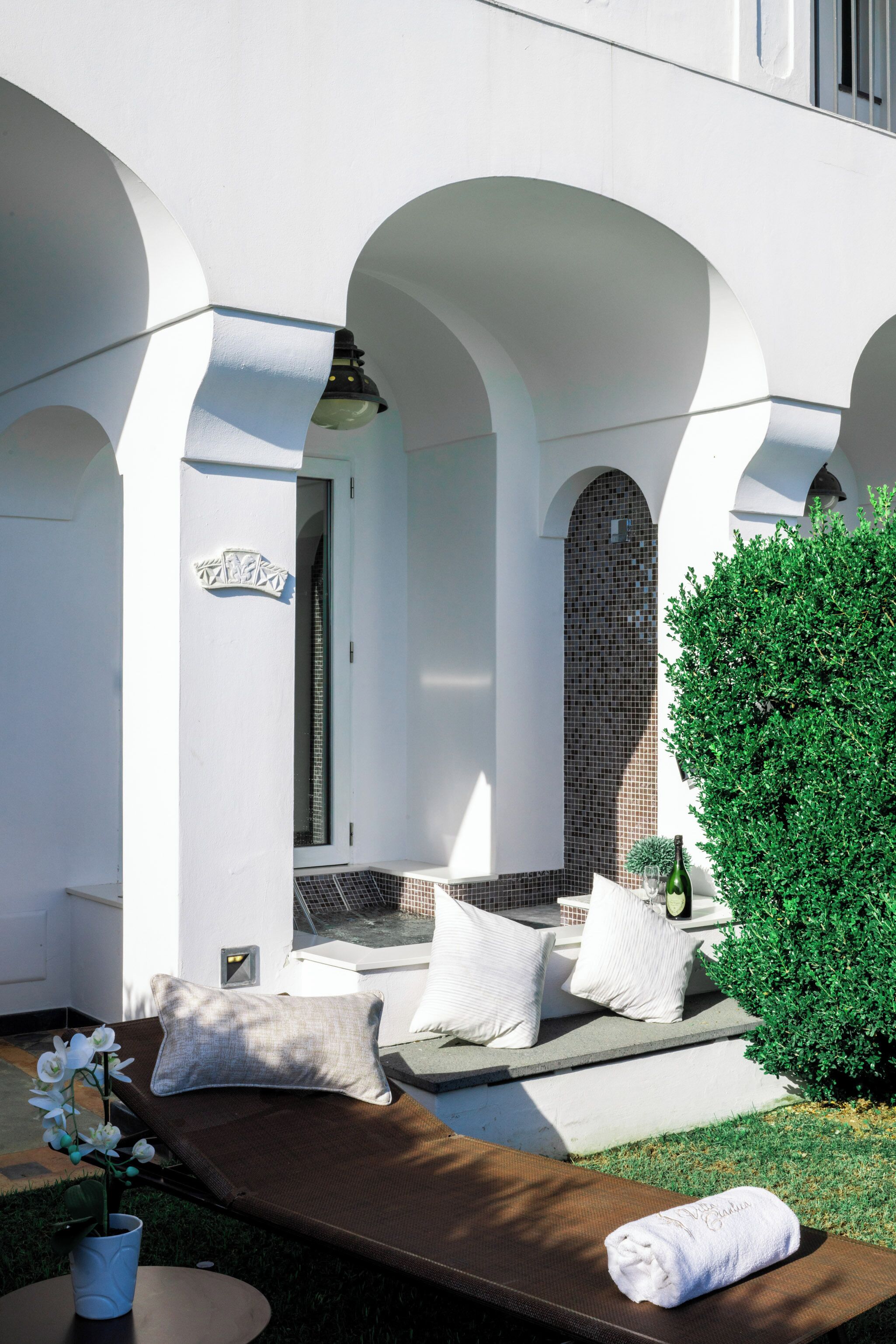 A sunlit outdoor seating area with a lounge chair, pillows, and a small table with a vase of flowers.