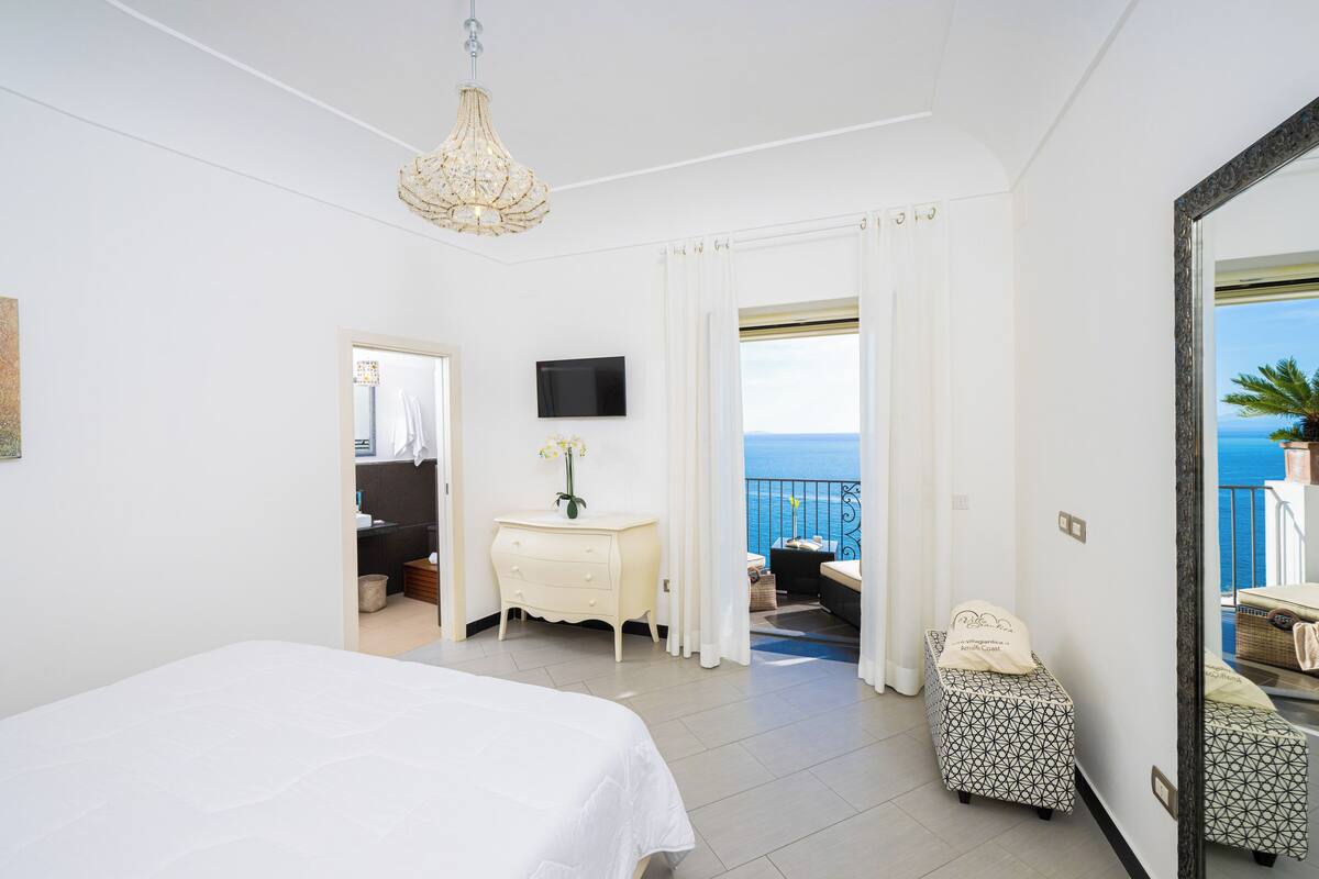 Een slaapkamer met een bed, een dressoir, een televisie en een balkon met uitzicht op zee.
