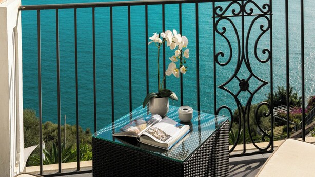Un balcón con vistas al mar, una mesa de mimbre con un florero y una revista.