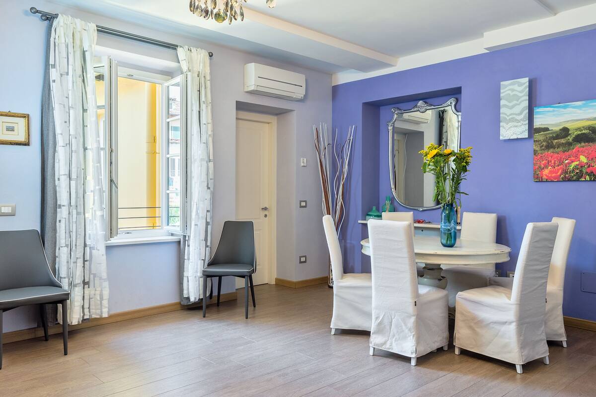 A dining area with a round table, white chairs, a vase with flowers, and a mirror on the wall.