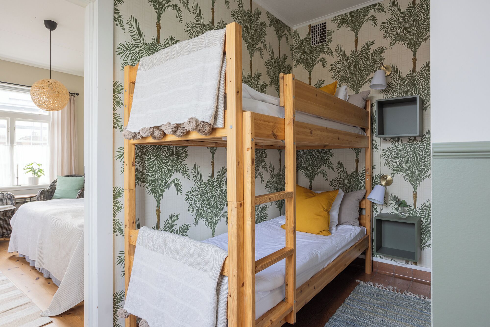 Une chambre avec un lit superposé, un papier peint aux motifs tropicaux, une table de chevet avec une lampe et une fenêtre avec des rideaux.
