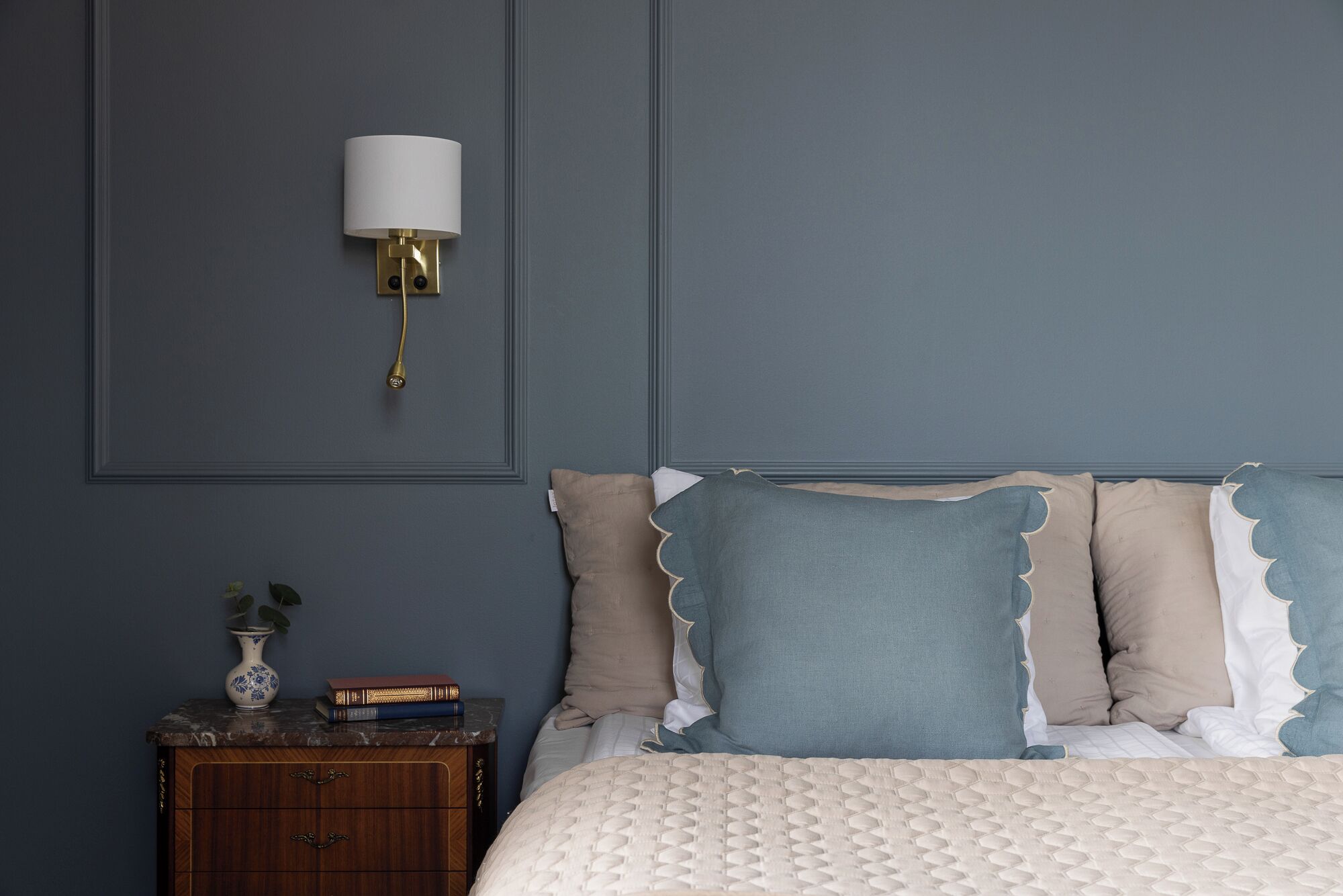 A bedroom with a bed, pillows, a nightstand, a vase, and a wall-mounted lamp.