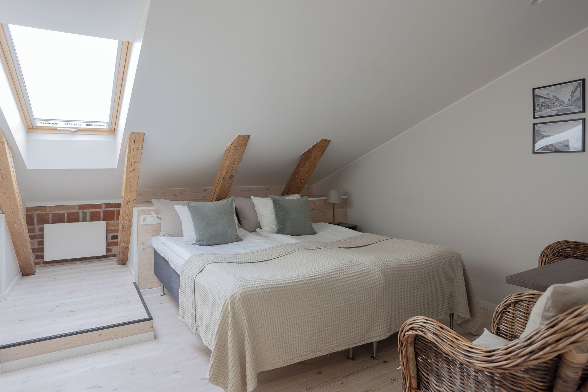 A bedroom with a sloped ceiling, a bed with a white and beige bedspread, a nightstand, a lamp, and a wicker chair.