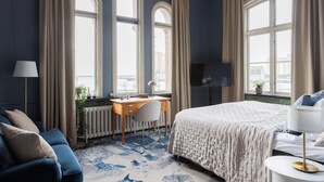 A bedroom with a large bed, a desk, and a chair. There are three large windows with curtains, a radiator, and a plant on the windowsill. - Stationen Bed & Breakfast (Ystad)