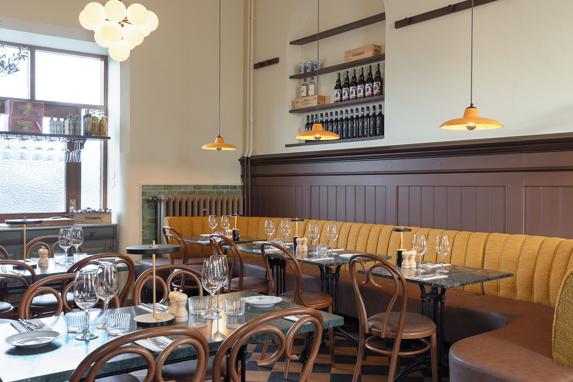 A restaurant interior with wooden booths, tables set for dining, hanging pendant lights, and shelves with bottles and glasses.