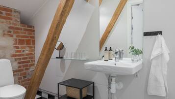 Un baño moderno con lavamanos, espejo y toallero blancos. Debajo del lavamanos, un estante negro con una cesta tejida. La habitación cuenta con vigas de madera a la vista y una pared de ladrillo.