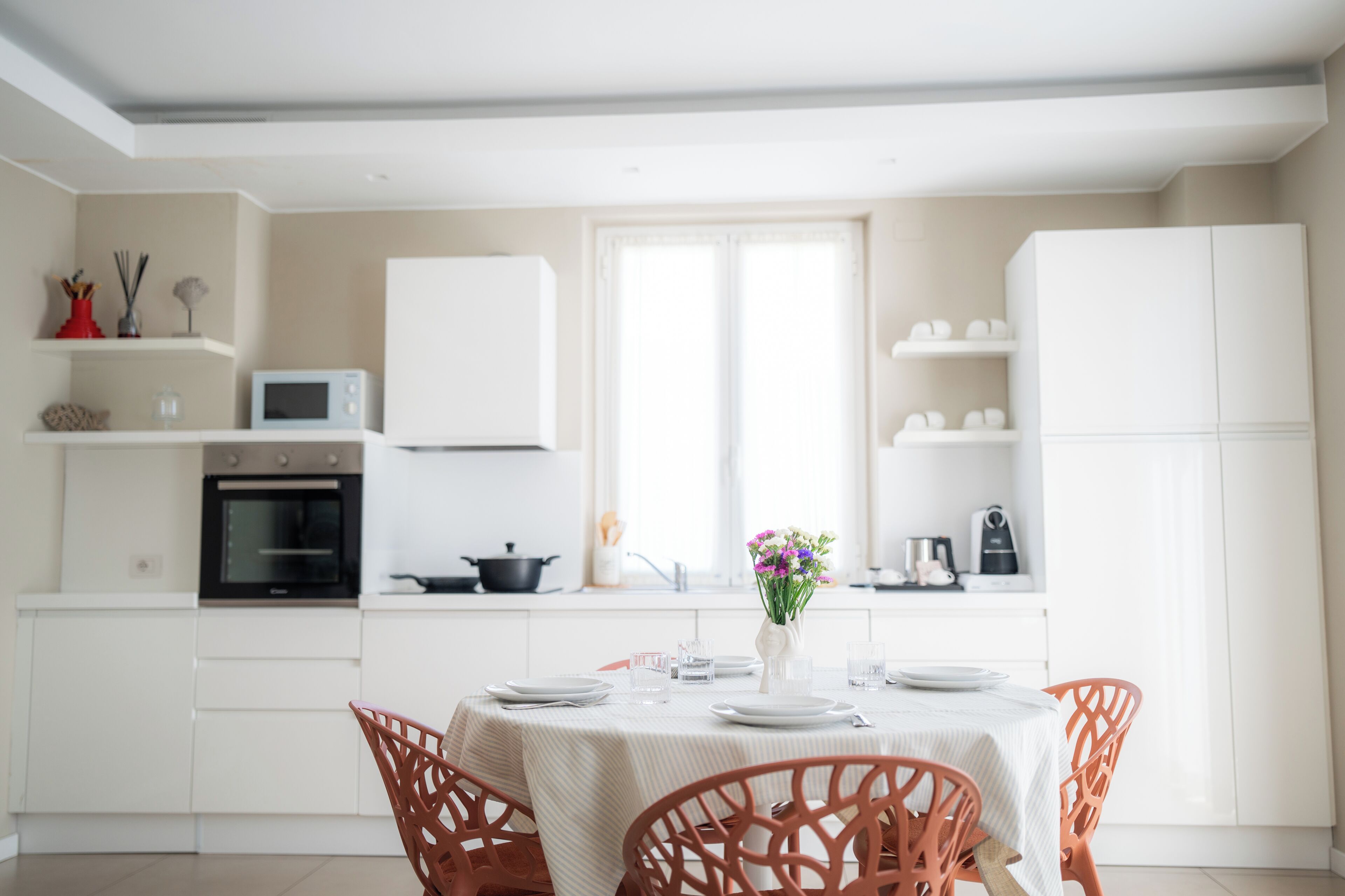 Uma cozinha moderna com armários brancos, uma mesa de jantar posta com pratos e um vaso de flores, e cadeiras laranja.
