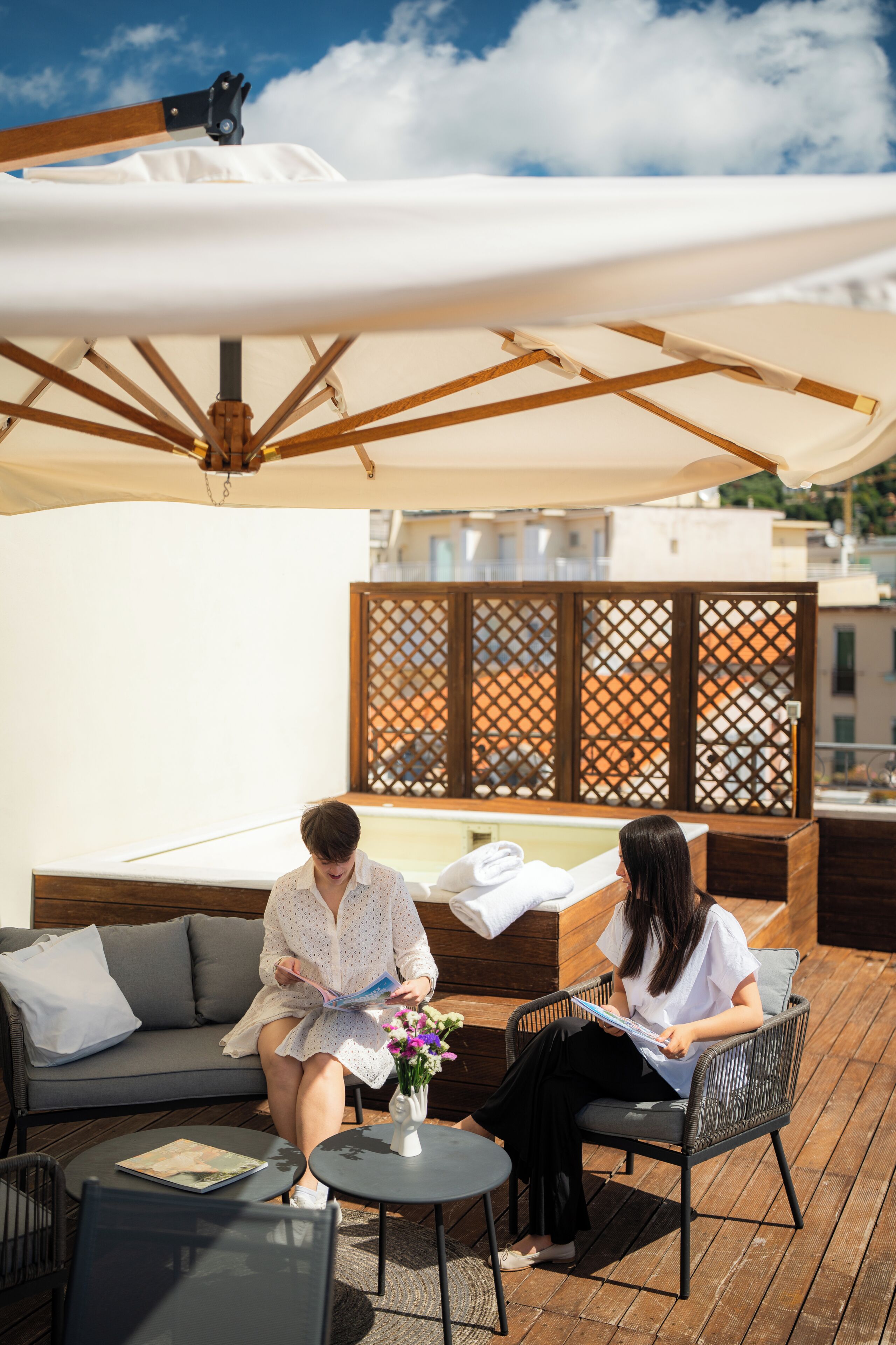 A covered outdoor seating area with a sofa, chairs, and a table, featuring a wooden deck and a view of buildings in the background.