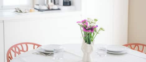 Una mesa de comedor dispuesta con platos, cubiertos y un jarrón de flores, con una cocina al fondo.