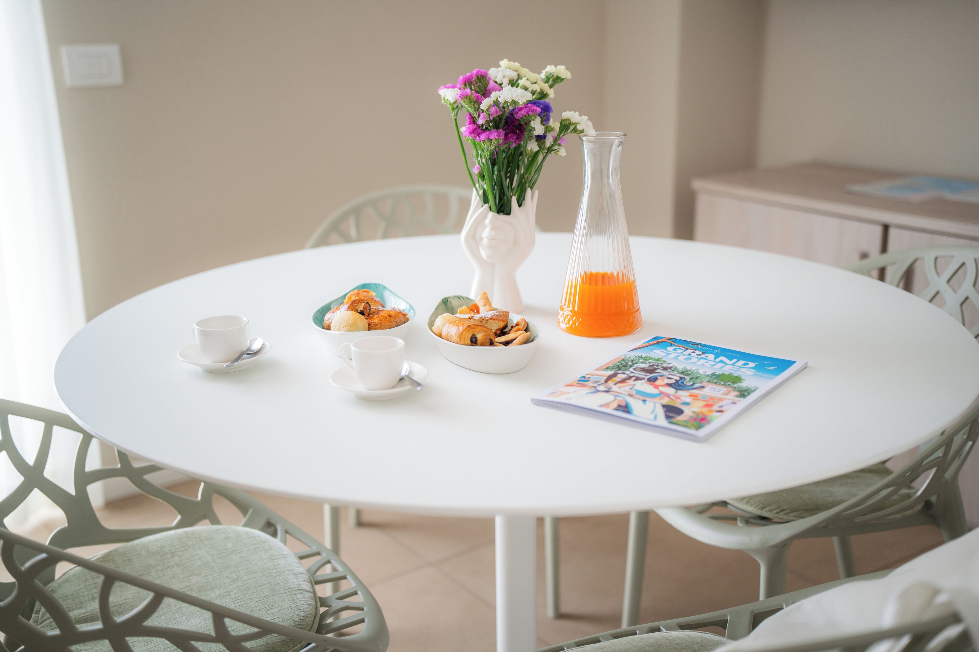 A round white table with a white vase, a glass pitcher, a bowl of pastries, two white cups, and a magazine.