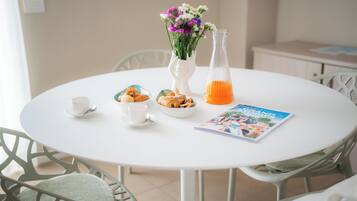 Une table ronde blanche, sur laquelle se trouvent un vase blanc, une carafe en verre, un bol rempli de viennoiseries, deux tasses blanches et un magazine.
