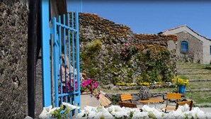Een landelijk stenen gebouw met een blauwe deur, een kleine tuin met bloemen en bankjes, en een vat.