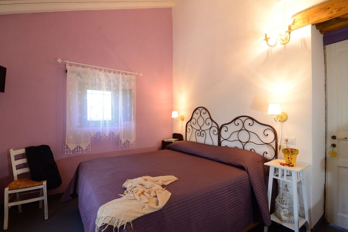 Een slaapkamer met een bed, een stoel, een nachtkastje, een lamp en een raam met kantgordijnen.