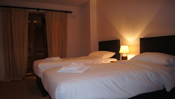 A hotel room with two beds, a nightstand with a lamp, and a door with curtains. - Castleforbes Holiday Apartment (Dublin)
