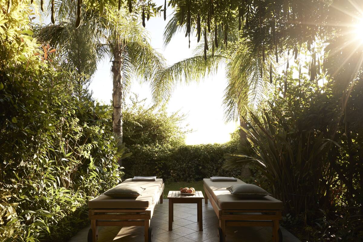 A serene outdoor relaxation area with two wooden lounge chairs and a small table, surrounded by lush greenery and palm trees, under a bright sun.