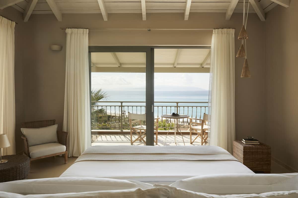 A bedroom with a large bed, a chair, a small table, and a view of the sea through a balcony.