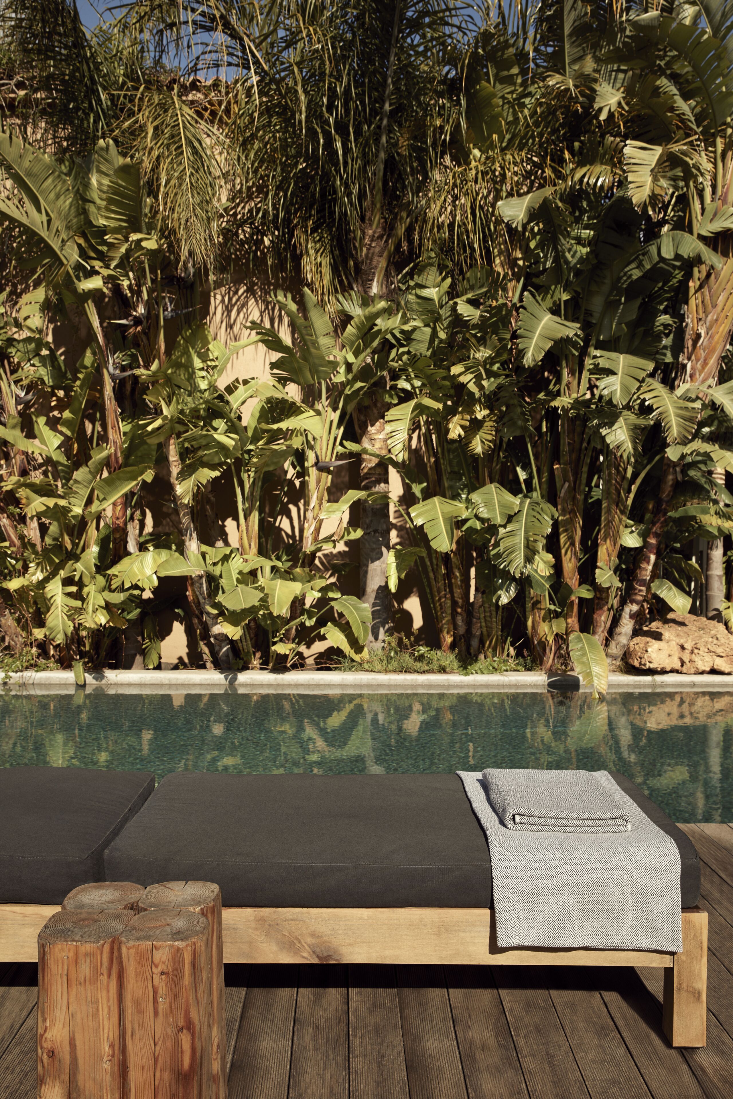 A wooden deck with a poolside seating area, a wooden stool, and a bench with a grey patterned throw.