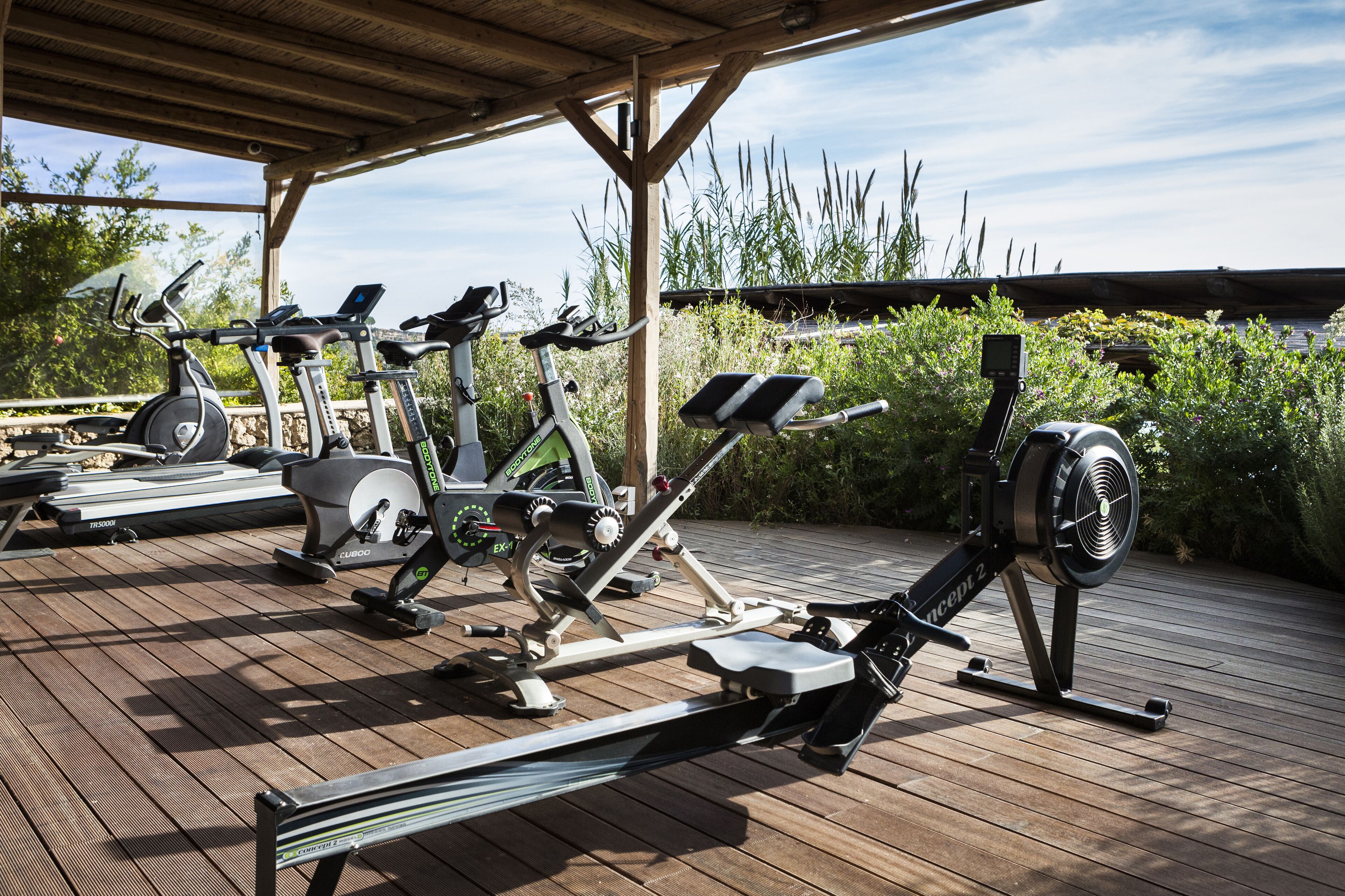 An outdoor gym with various exercise equipment, including treadmills, stationary bikes, and a rowing machine, under a wooden shelter.