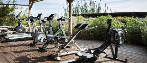 An outdoor gym with various exercise equipment, including treadmills, stationary bikes, and a rowing machine, under a wooden shelter.