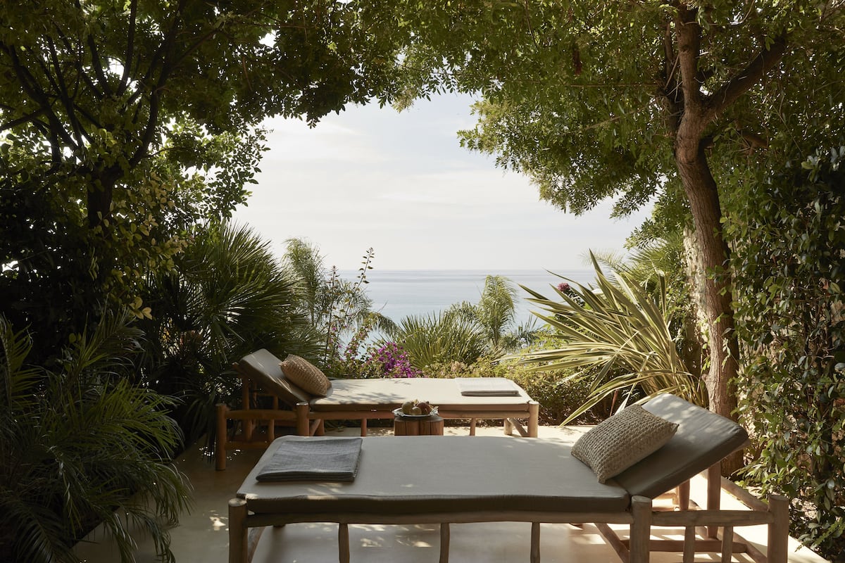 A sunlit outdoor seating area with two wooden lounge chairs and a table, surrounded by lush greenery and overlooking a body of water.
