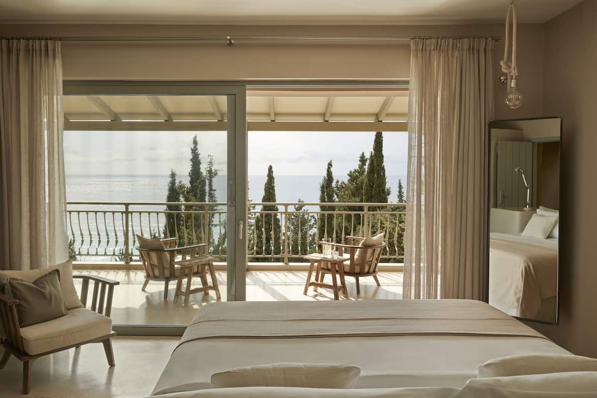 A bedroom with a large bed, a balcony with a table and chairs, and a view of the sea.