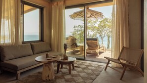 Une pièce comprenant un canapé, deux petites tables rondes, un fauteuil en osier et une grande porte-fenêtre coulissante donnant sur un espace extérieur avec vue sur la mer et un parasol de plage.