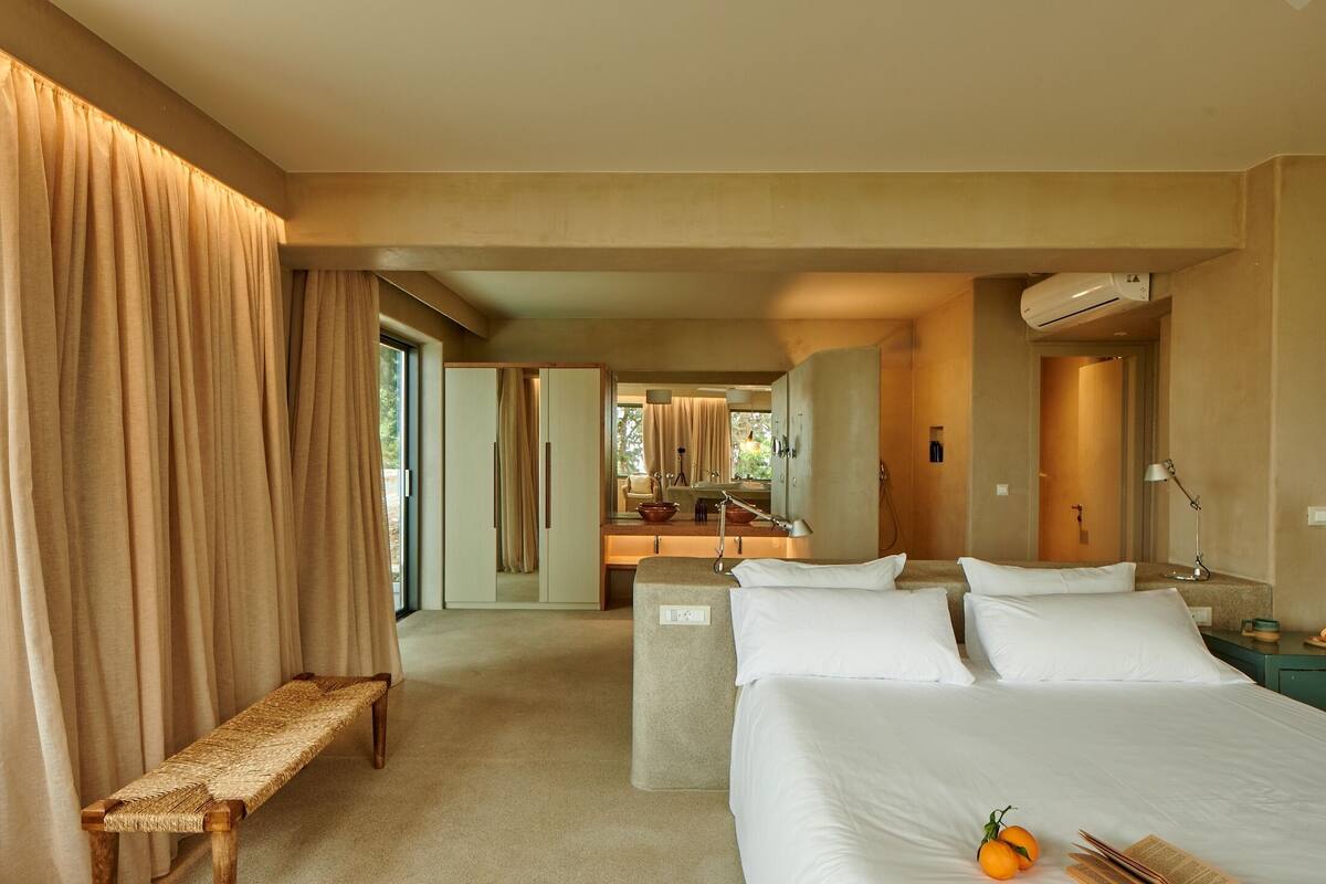 A hotel room with a large bed, a bench, a desk with a mirror, and an air conditioning unit.
