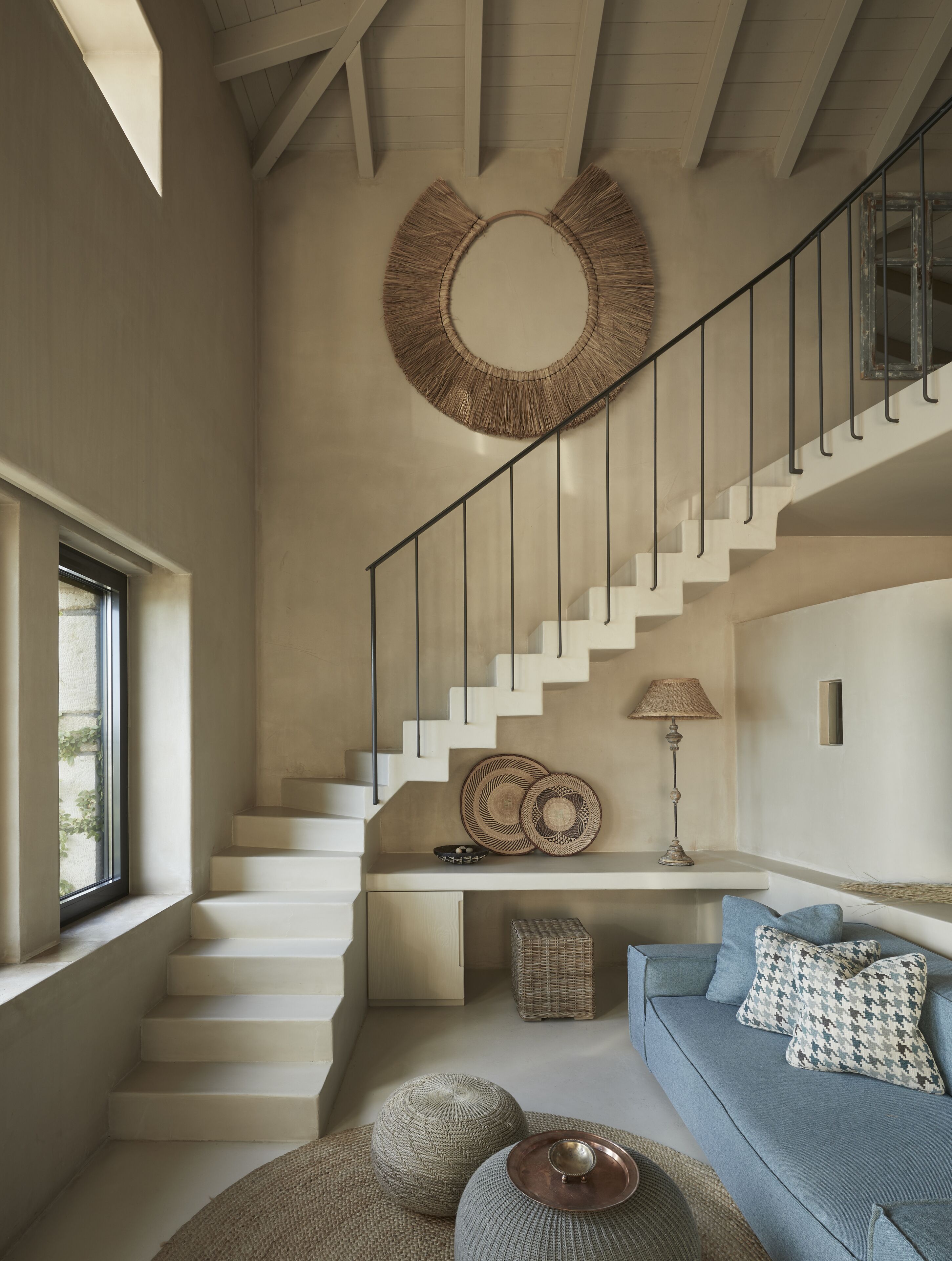 Un vaste espace de vie comprenant un escalier, une grande tapisserie murale tissée et un canapé bleu agrémenté de coussins à motifs.
