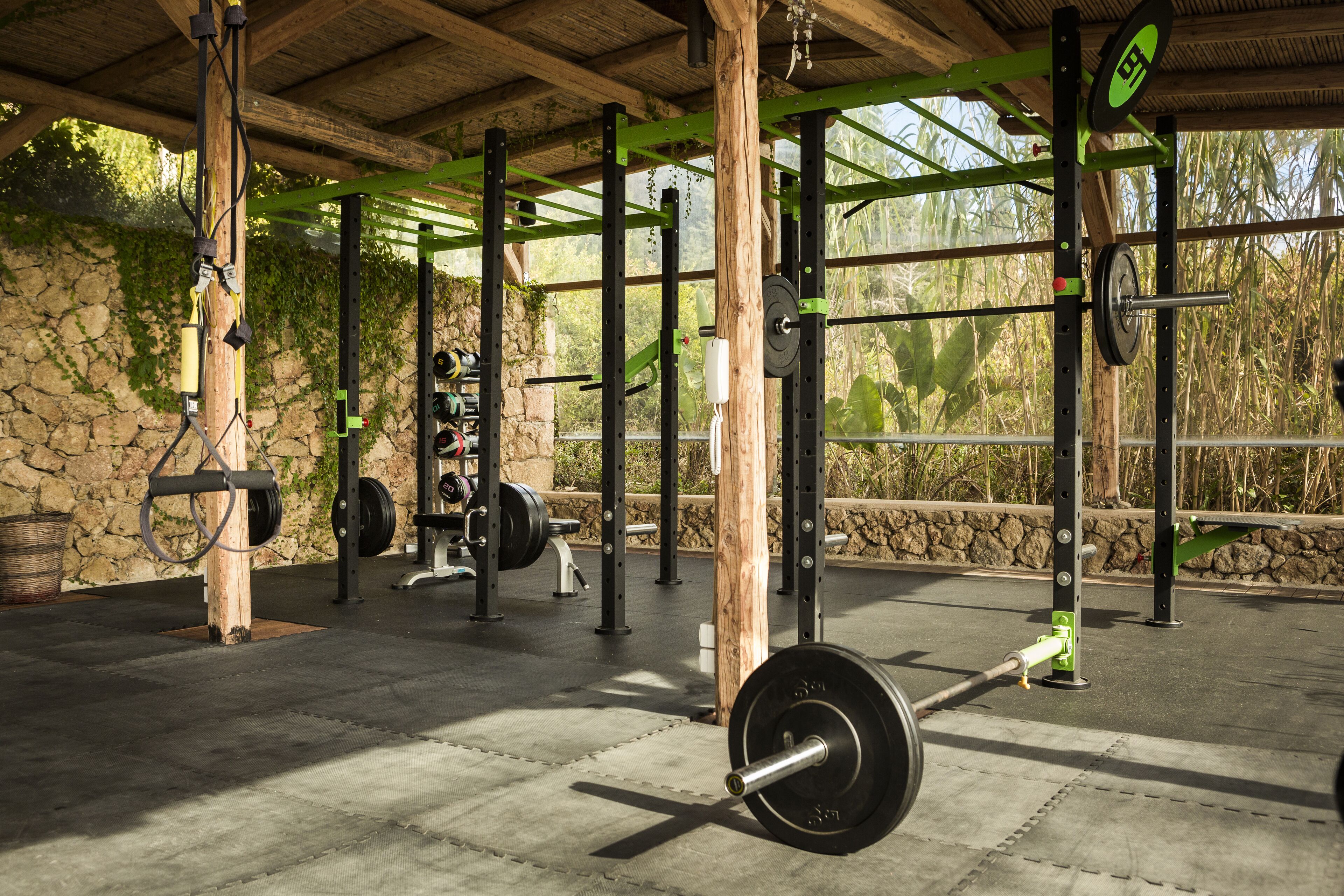 A covered outdoor gym with weightlifting equipment and a stone wall backdrop.
