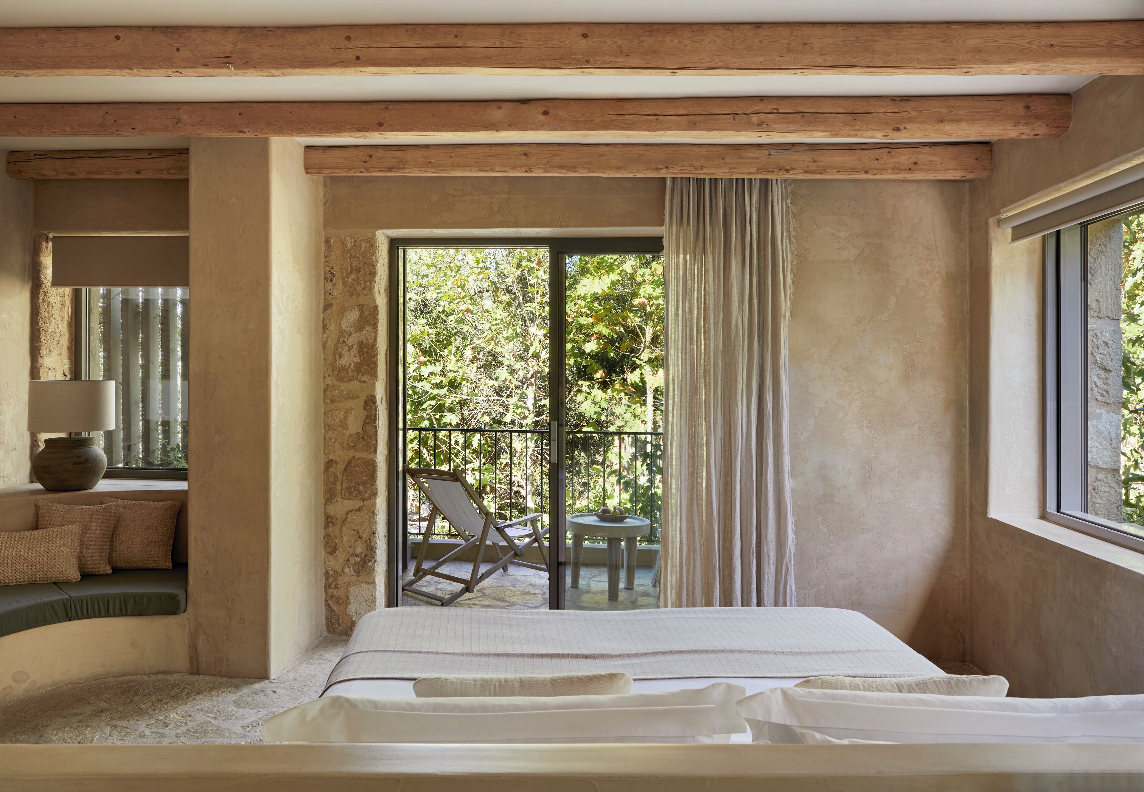 A bedroom with a large bed, a view of the outdoors through a sliding door, a chair, and a table outside.