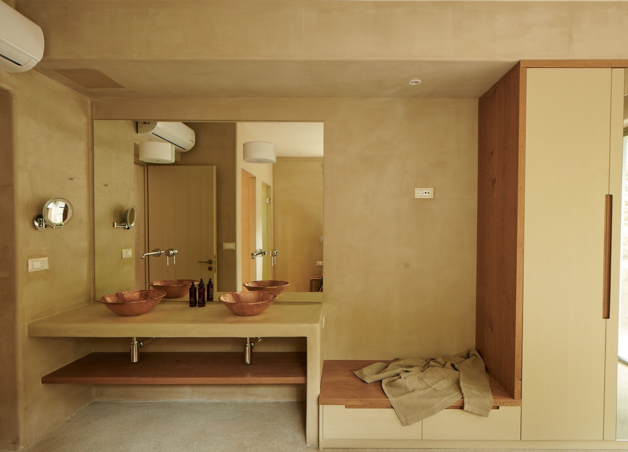 A modern bathroom with a large mirror, two copper sinks, and a wooden bench with a towel.
