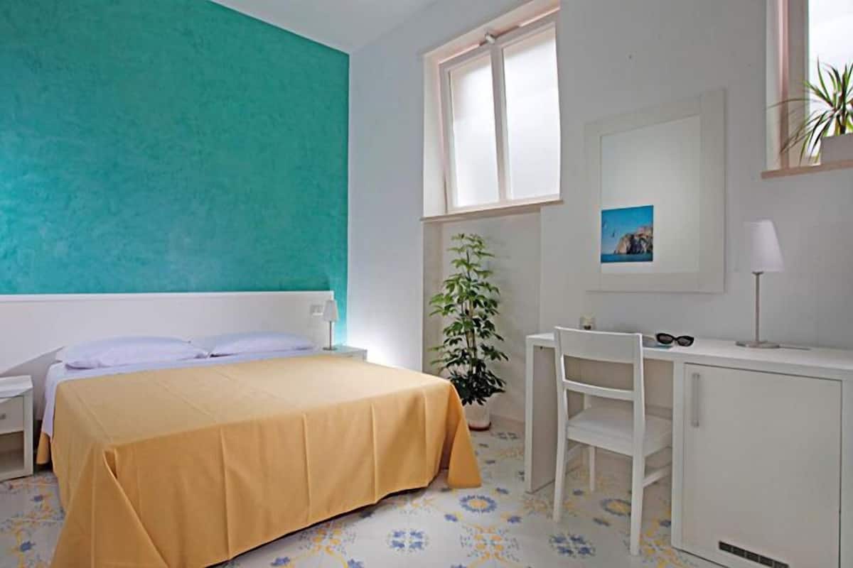 A bedroom with a bed, a desk, a chair, a potted plant, a window, and a wall with a picture.