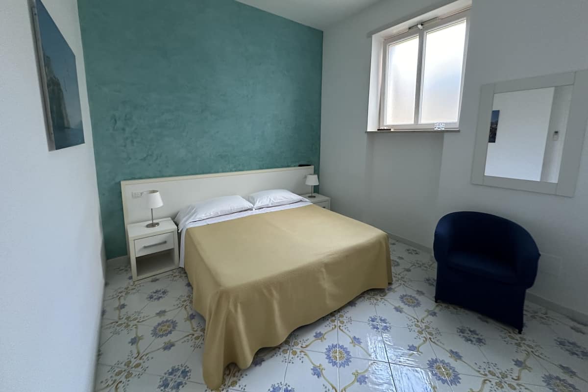 A bedroom with a bed, a nightstand, a chair, a window, and a wall with a picture.