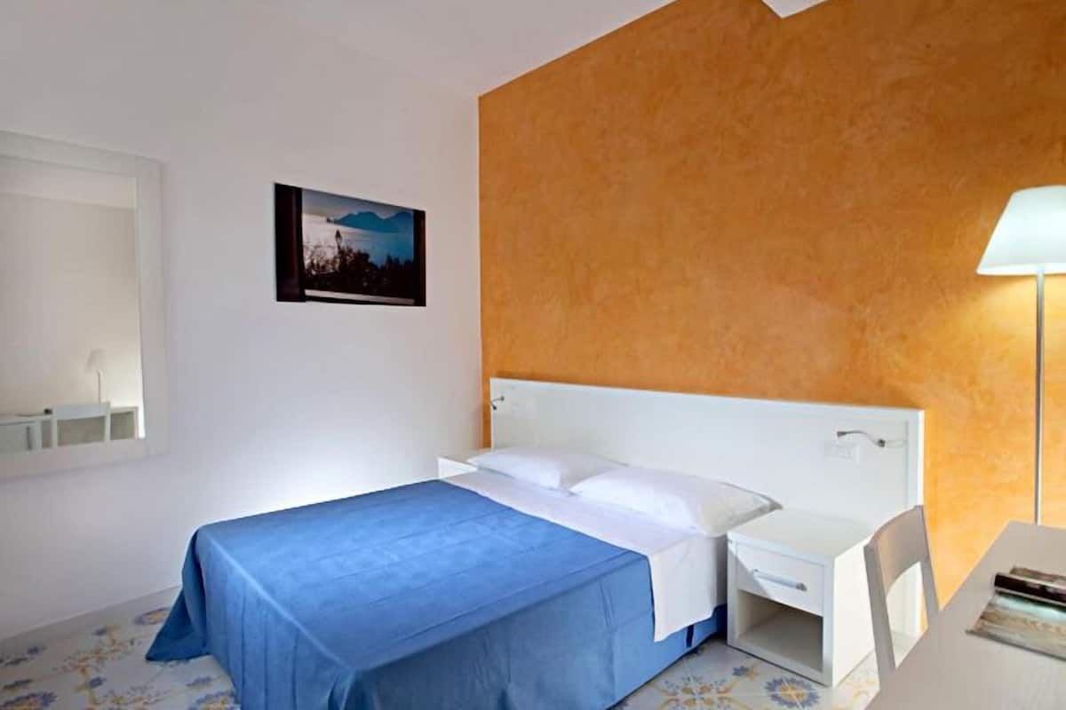 A hotel room with a bed, a desk, a chair, a lamp, and a picture on the wall.