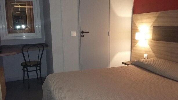 A bedroom with a bed, a bedside table, a chair, a wardrobe, and a window. - Hotel du Triangle (Talange)