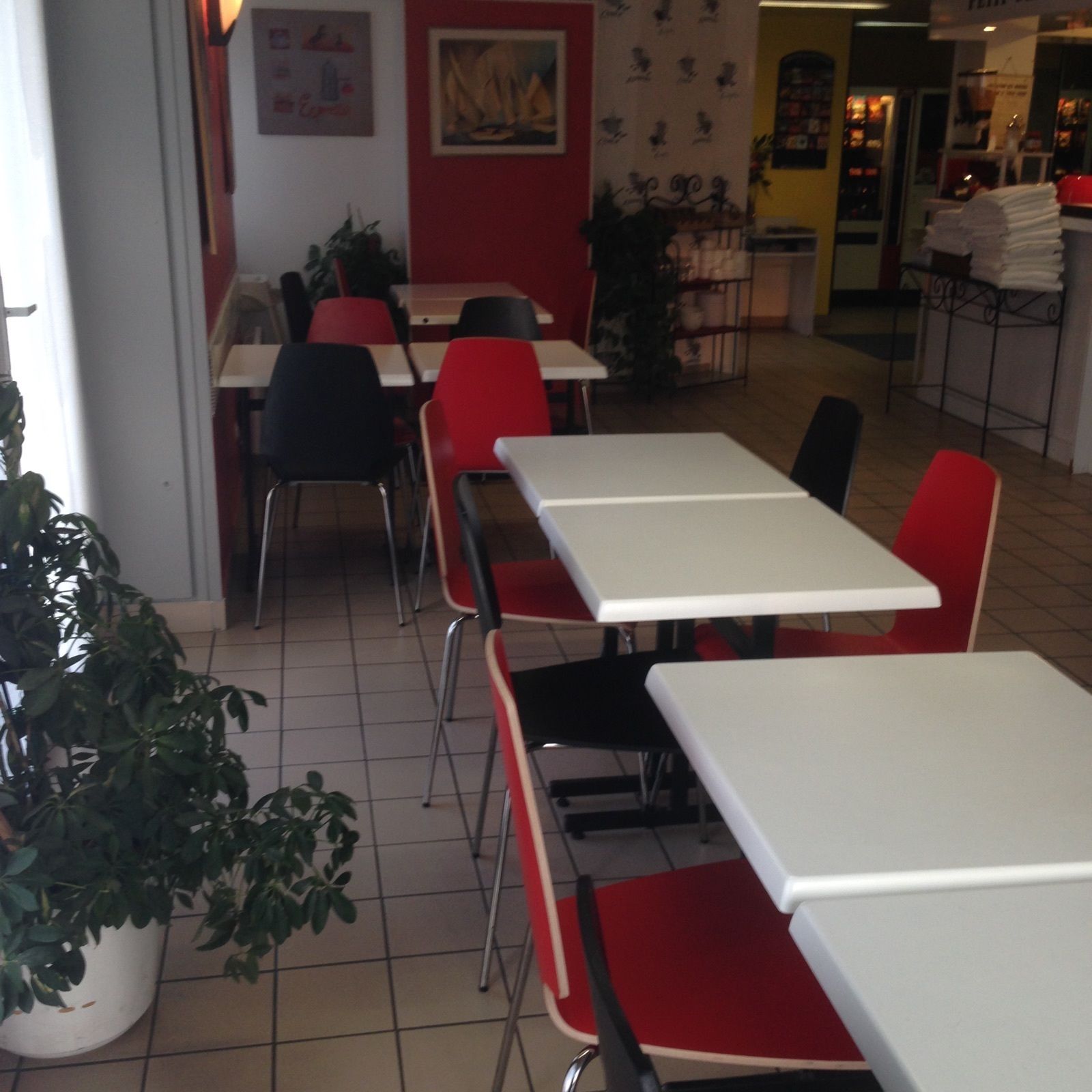 A restaurant interior with white tables and red chairs, potted plants, and tiled flooring.