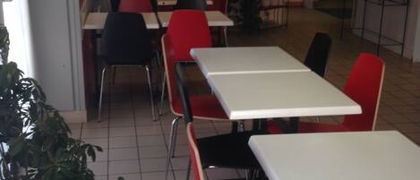 A restaurant interior with white tables and red chairs, potted plants, and tiled flooring.