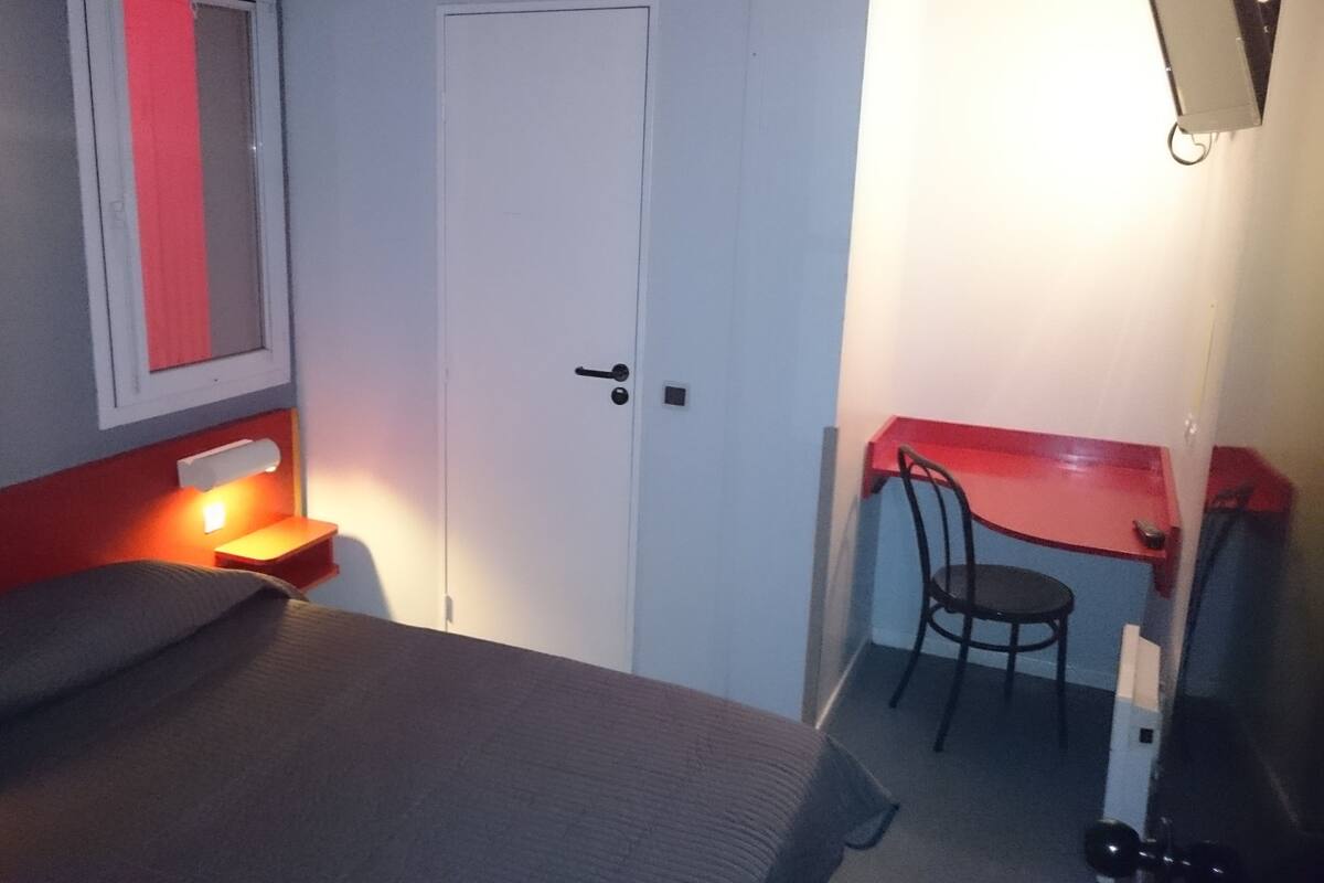 A small, compact room with a bed, a desk, and a chair. There is a door with a handle and a light switch on the wall.