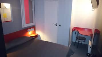 Ein kleiner, kompakter Raum mit einem Bett, einem Schreibtisch und einem Stuhl. An der Wand befindet sich eine Tür mit Griff und ein Lichtschalter.