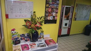 A reception area with a whiteboard displaying a floor plan, a flower arrangement, and various brochures and magazines on a table. - Hotel du Triangle (Talange)
